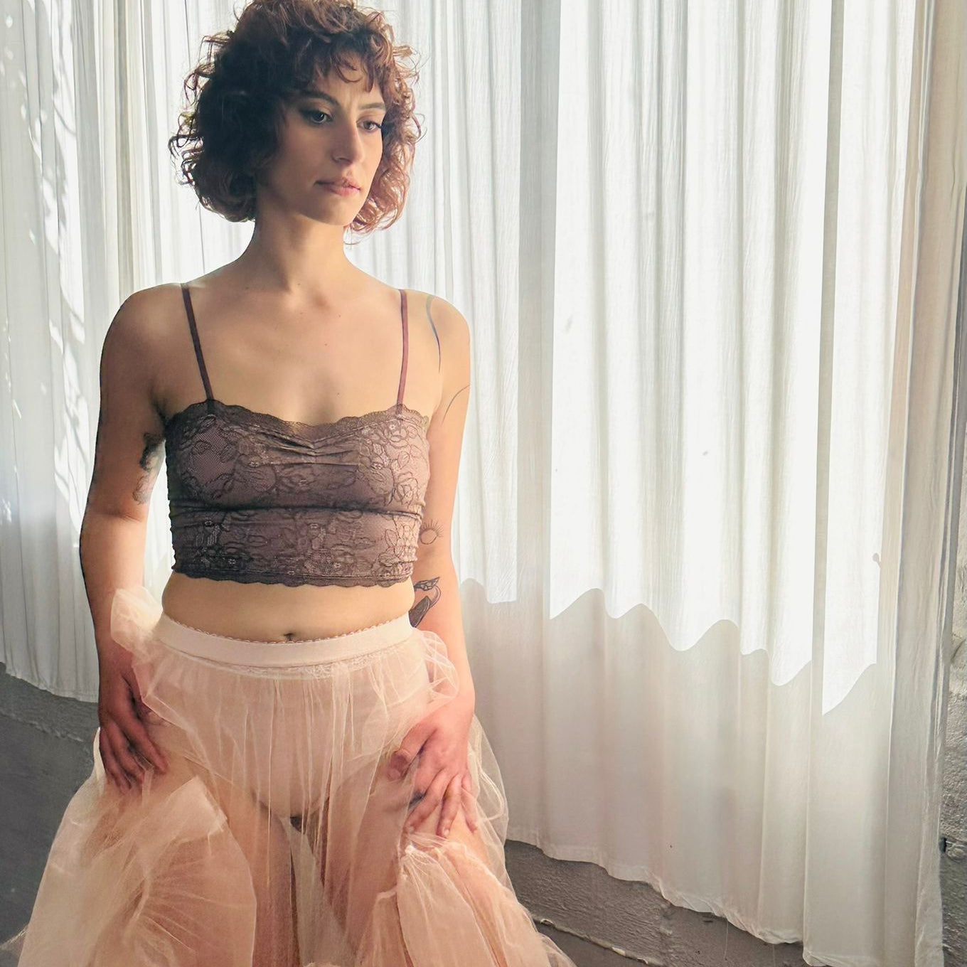 Woman wearing a sheer top and tulle skirt in front of white curtains.