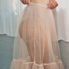 Person wearing a sheer peach skirt and bra against a white curtain background